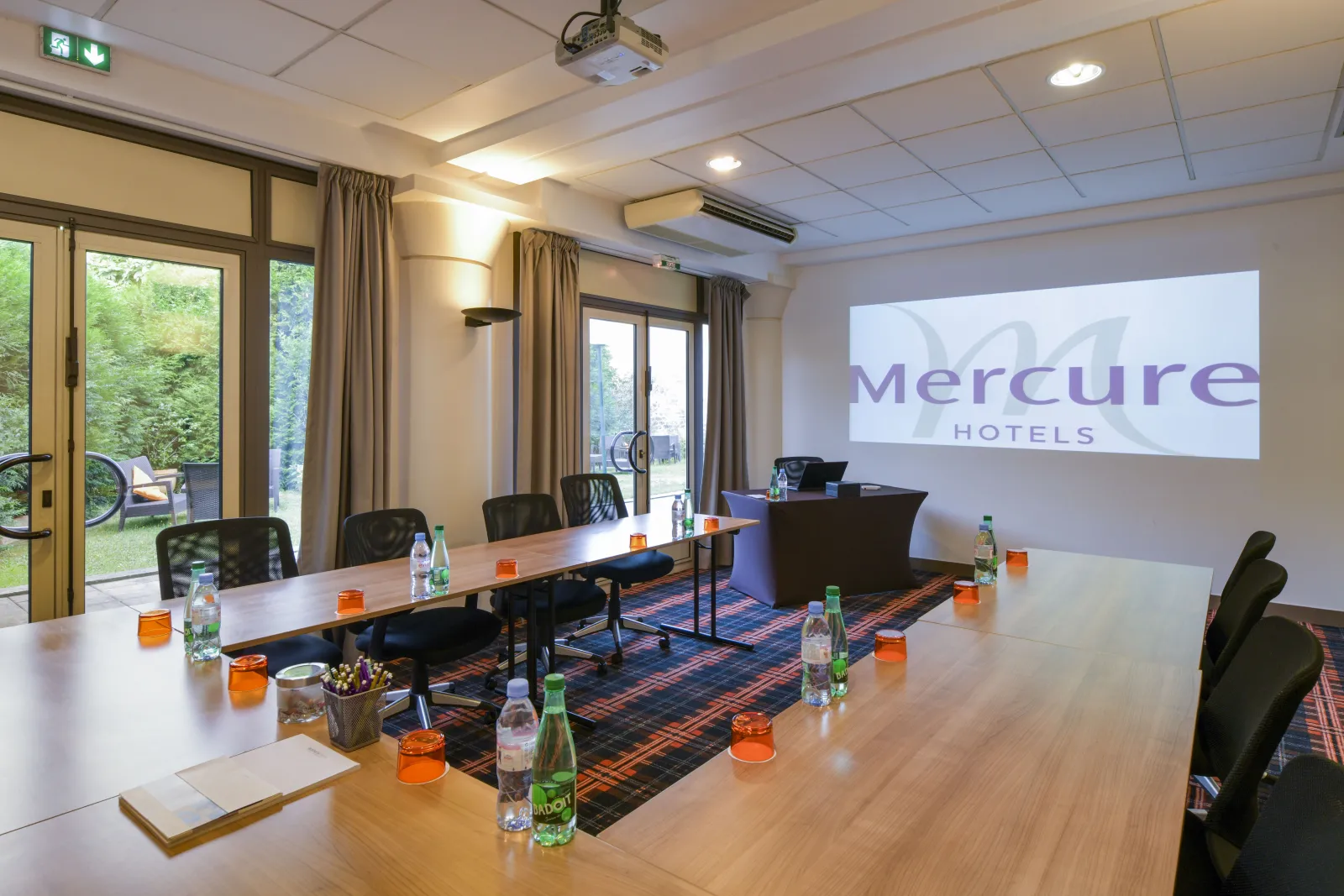 Modern conference room at Mercure Bordeaux Château Chartrons with long wooden tables, black chairs, water bottles, projection screen displaying Mercure Hotels logo, and glass doors opening to a garden area.