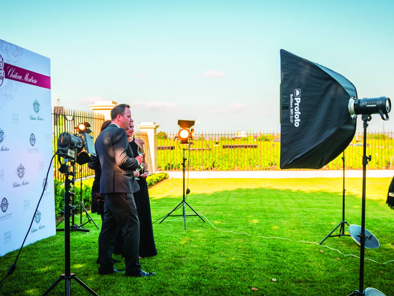 A man in a dark suit speaks at a microphone during an outdoor event in a sunflower field, with professional lighting equipment and a branded backdrop visible.