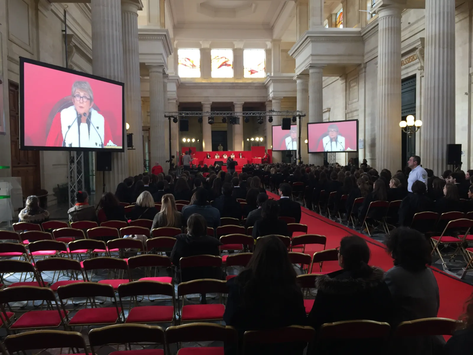 Large conference hall in Bordeaux with classical architecture, red carpet and seating, displaying a speaker on pink-background screens, with attendees seated throughout the ornate space featuring tall columns and natural light from upper windows.