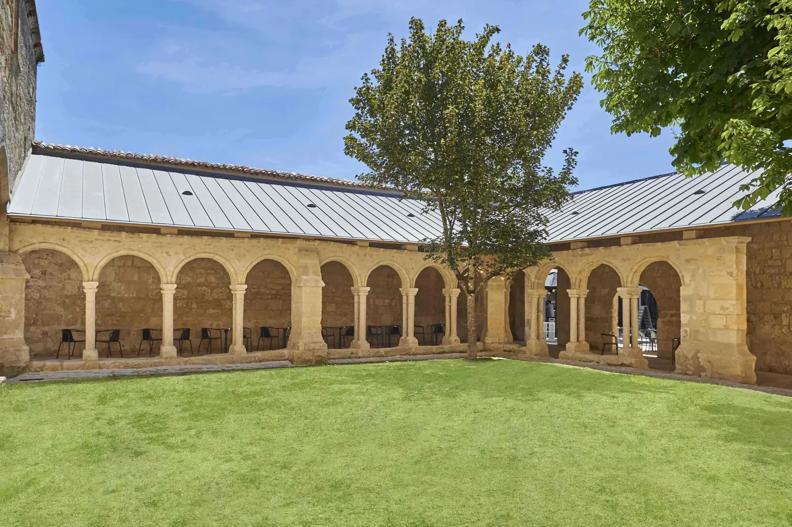 Cloister courtyard of Les Cordeliers in Bordeaux featuring Romanesque arches, manicured lawn, and modern metal roof, with medieval stone architecture surrounding the space.