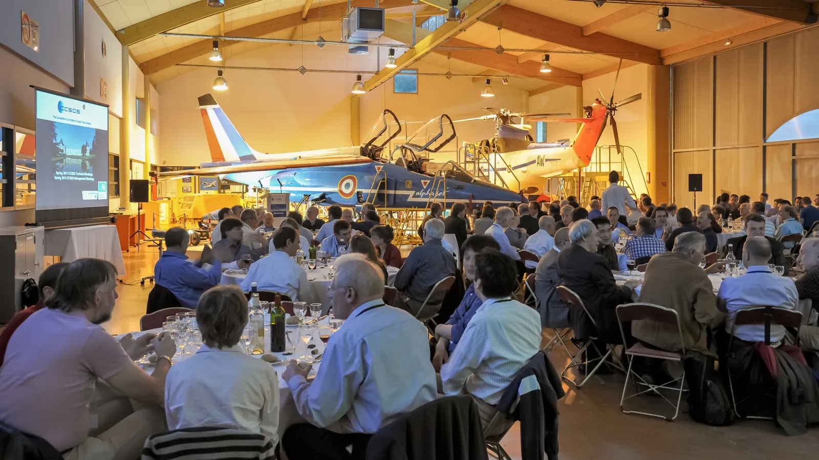 A formal dinner event held inside a hangar at Aérocampus Aquitaine in Bordeaux, with dozens of guests seated at tables in front of a restored military aircraft on display. A presentation screen is visible on the left side of the venue.