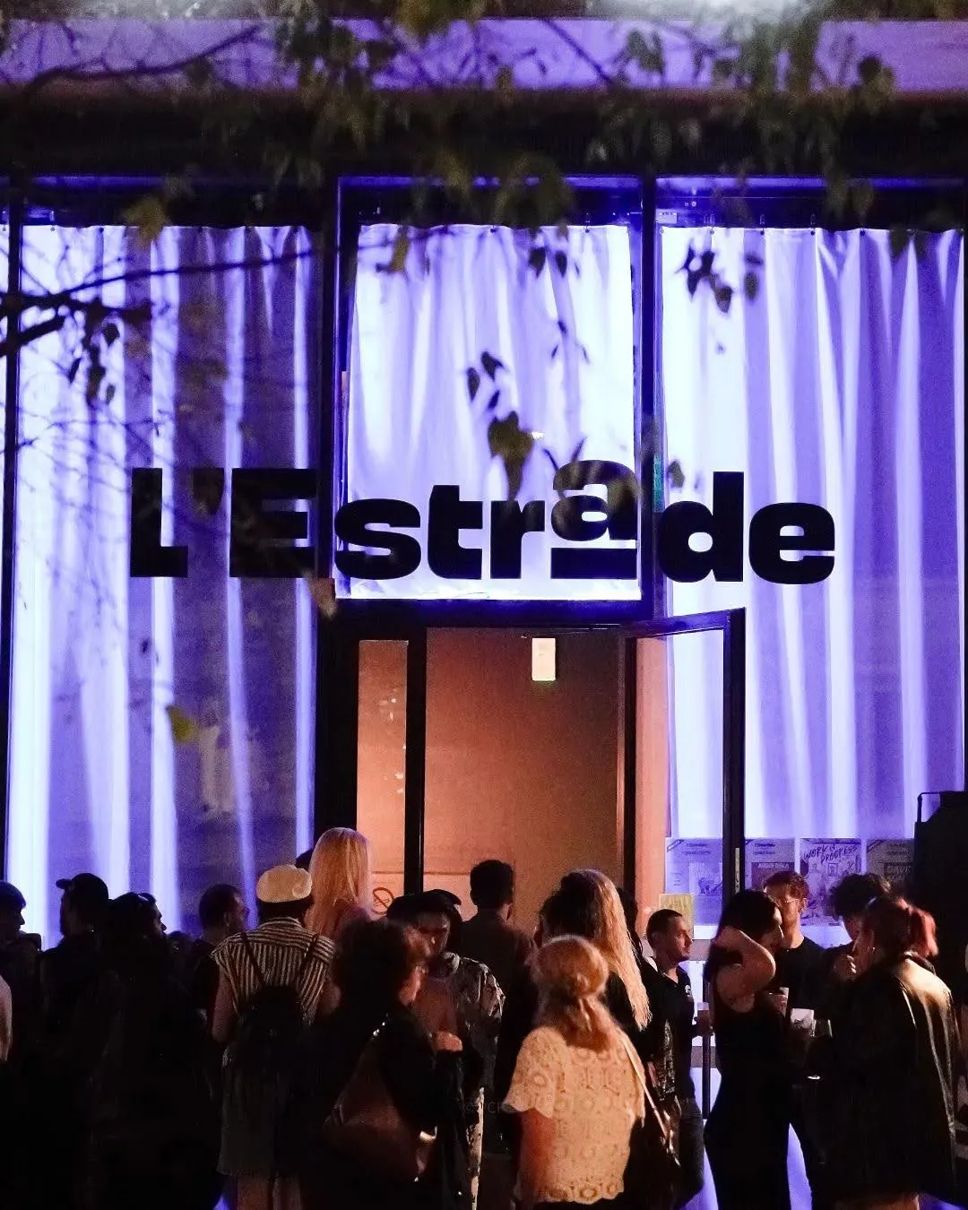Nighttime exterior view of L'Estrade venue in Bordeaux with illuminated purple and white striped facade, crowds of people gathered at the entrance