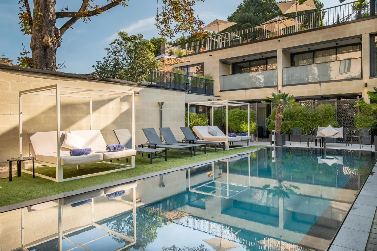 Luxury pool area at Palais Gallien Hotel & Spa in Bordeaux, featuring a modern swimming pool with sun loungers, white canopy beds, manicured lawn, and contemporary architecture with terraces overlooking the resort.