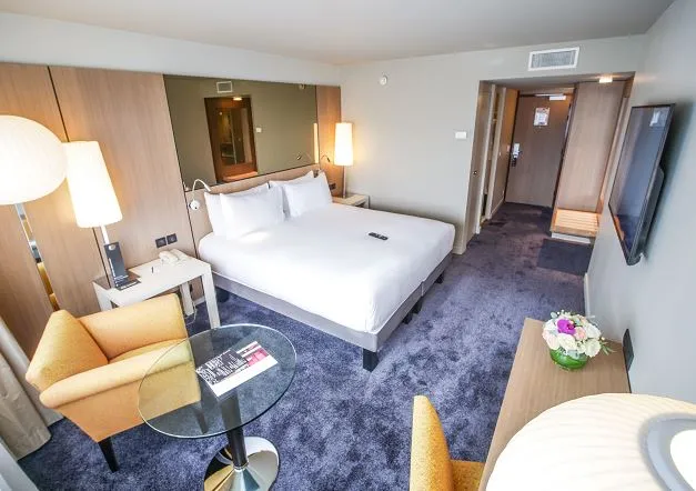 Modern hotel room at Pullman Bordeaux Lac featuring a king-size bed with white linens, comfortable seating area with yellow armchair, dark blue carpet, ambient lighting, and contemporary furnishings