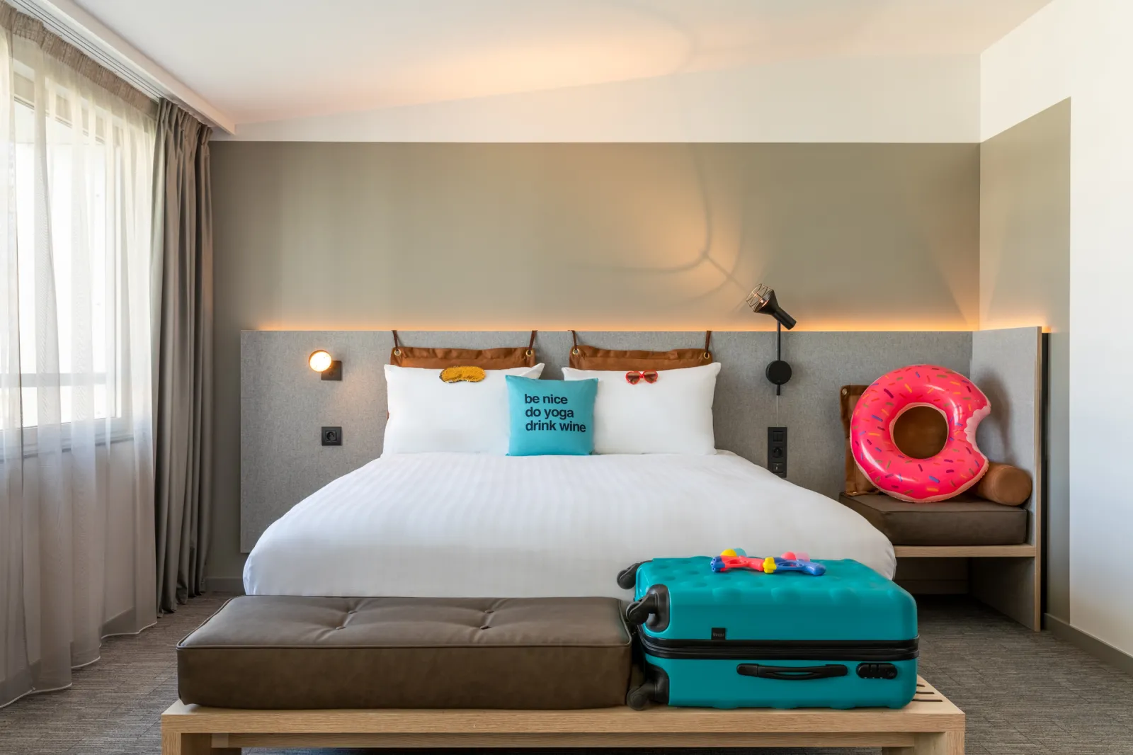 Modern hotel bedroom at Moxy Bordeaux featuring a queen bed with gray upholstered headboard, turquoise pillow with motivational text, white bedding, ambient warm lighting, colorful travel accessories including a pink inflatable donut and turquoise suitcas
