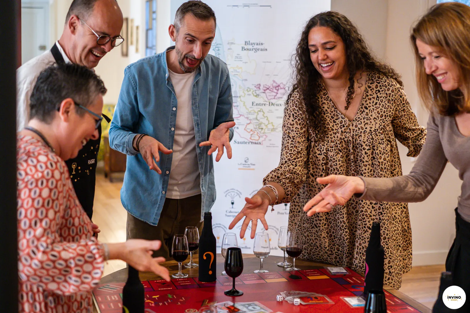Five people gathered around a table with wine glasses and tasting cards, smiling and engaging in a wine tasting activity. A wine region map is visible on the wall in the background.