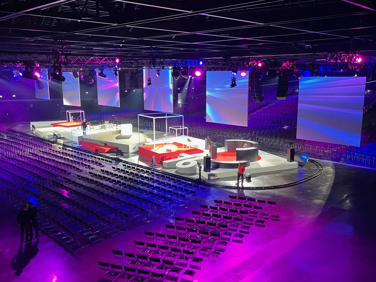 Modern indoor arena with purple and blue lighting, red and white modular stage setup in the center, rows of black seating, large video screens, and professional rigging equipment in Bordeaux.