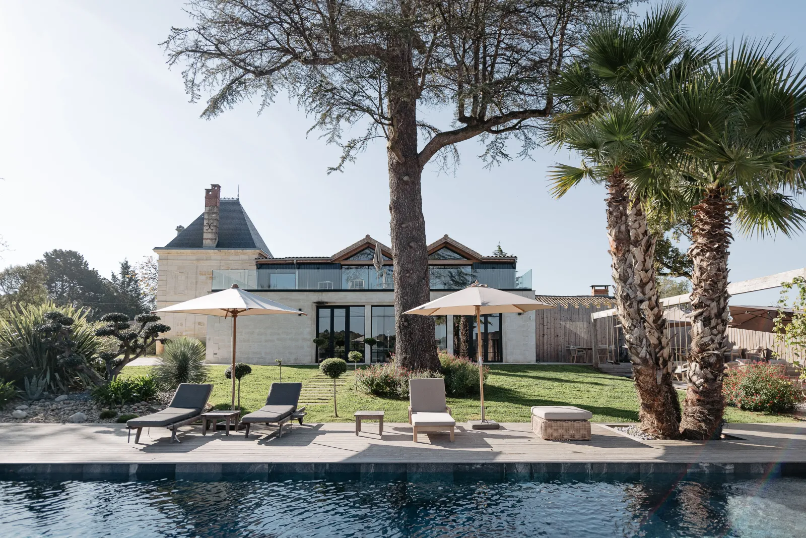 Modern luxury estate with pool, featuring a renovated château tower, contemporary white architecture, palm trees, loungers, and manicured gardens under clear sky