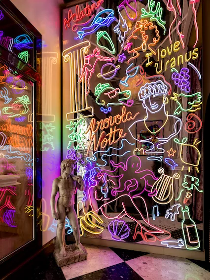 Vibrant neon sign installation at Osteria Palatino in Bordeaux, featuring colorful glowing tubes with musical and artistic motifs including instruments, grapes, and Italian text against a dark background.