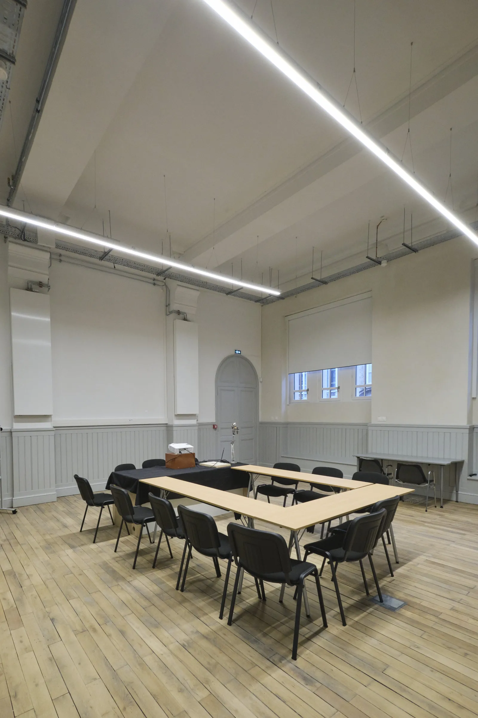 Modern meeting room with U-shaped wooden table, black chairs, linear ceiling lights, white walls with paneling, and large windows at ManuCo in Bordeaux.