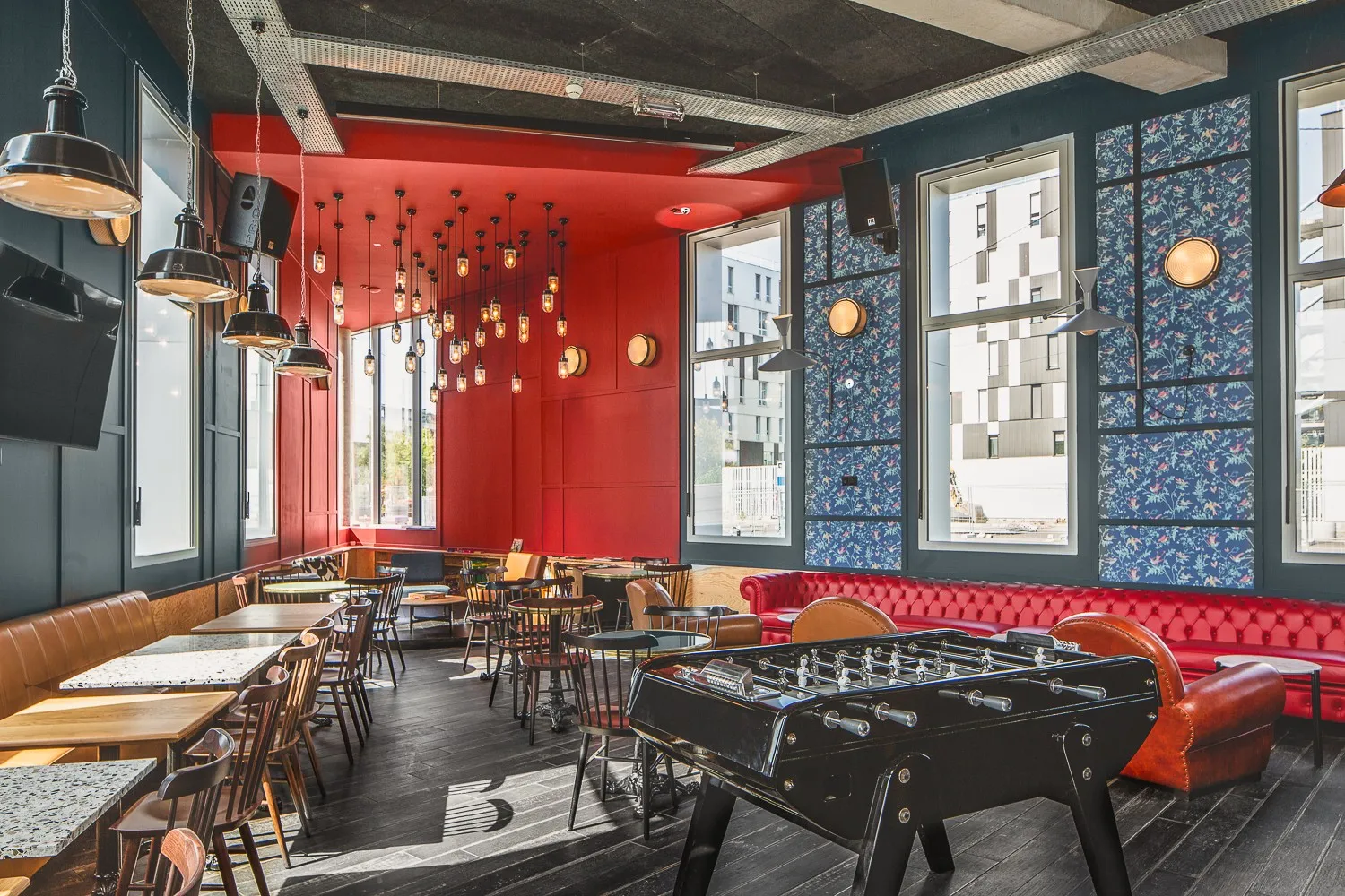 Modern hotel lounge at Eklo Bordeaux featuring a red accent wall with pendant lights, leather seating, foosball table, and blue patterned wallpaper with city views through large windows
