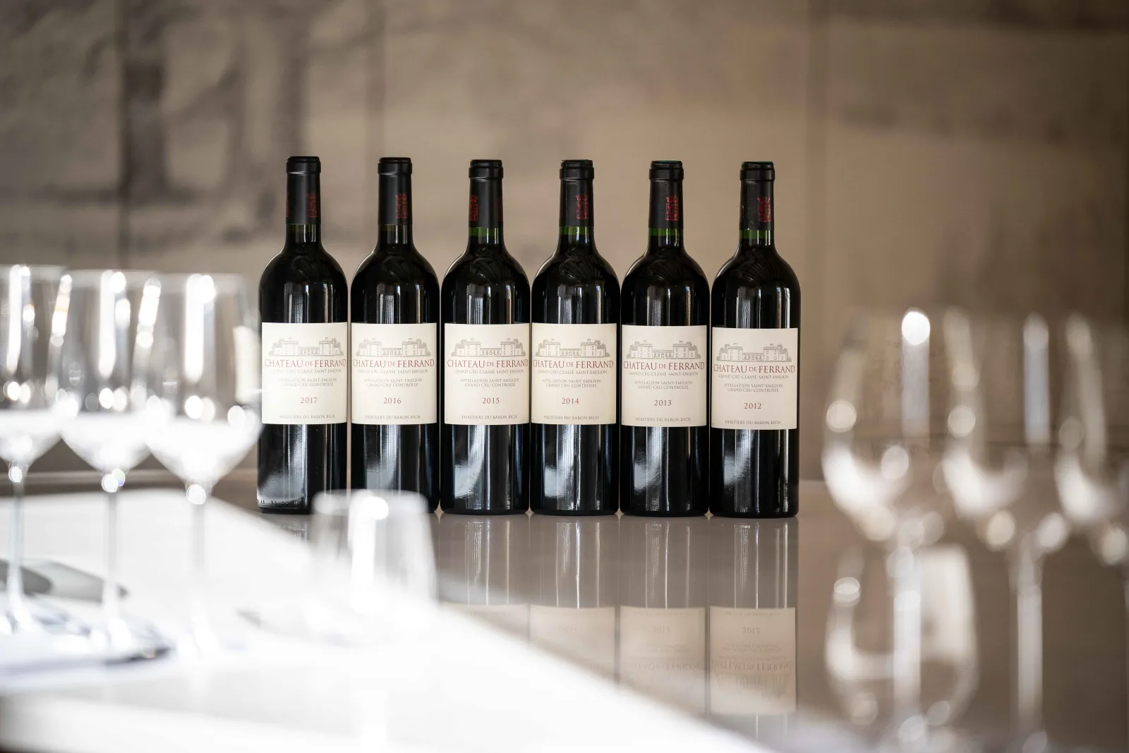 Six bottles of Château de Ferrand wine from vintages 2012 to 2017 displayed in a row on a white table with wine glasses in the background, in an elegant dining setting.