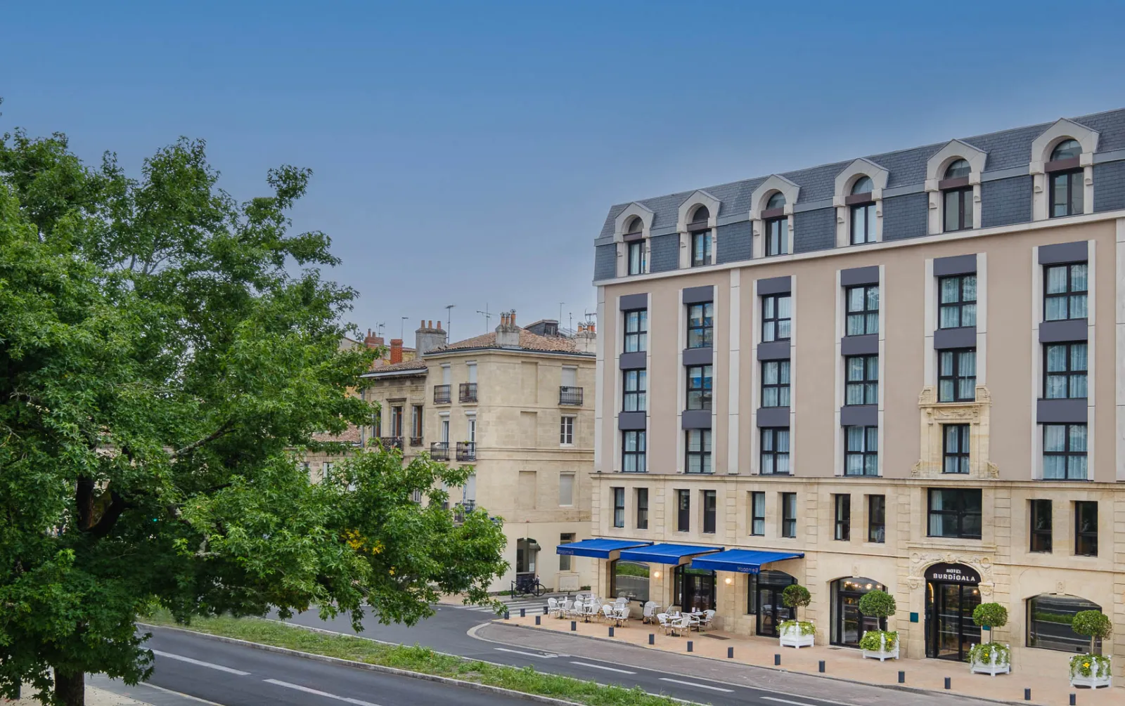 Exterior view of Hôtel Burdigala by Inwood Hotels in Bordeaux, a beige and blue Haussmann-style building with mansard roof, ground-floor café with blue awning, and tree-lined street.