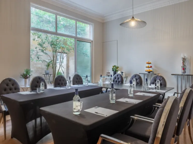 Modern conference room with U-shaped arrangement of black tables, gray upholstered chairs, water bottles, and notepads, featuring large windows overlooking a garden and a contemporary pendant light.