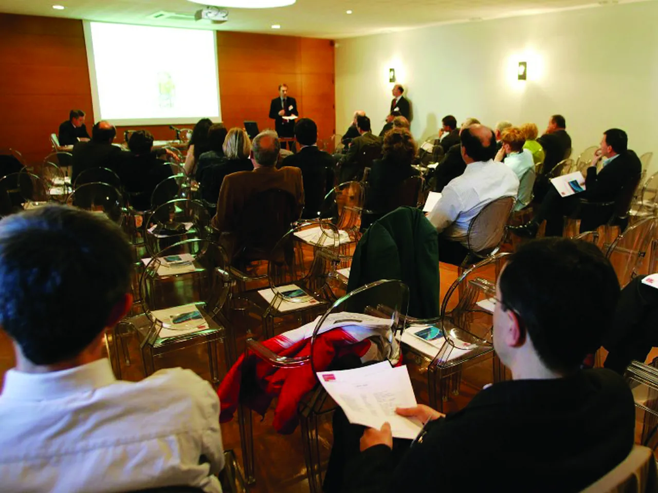 Business conference or seminar in Bordeaux with attendees seated in a meeting room facing two speakers near a projection screen, with an orange accent wall visible in the background.
