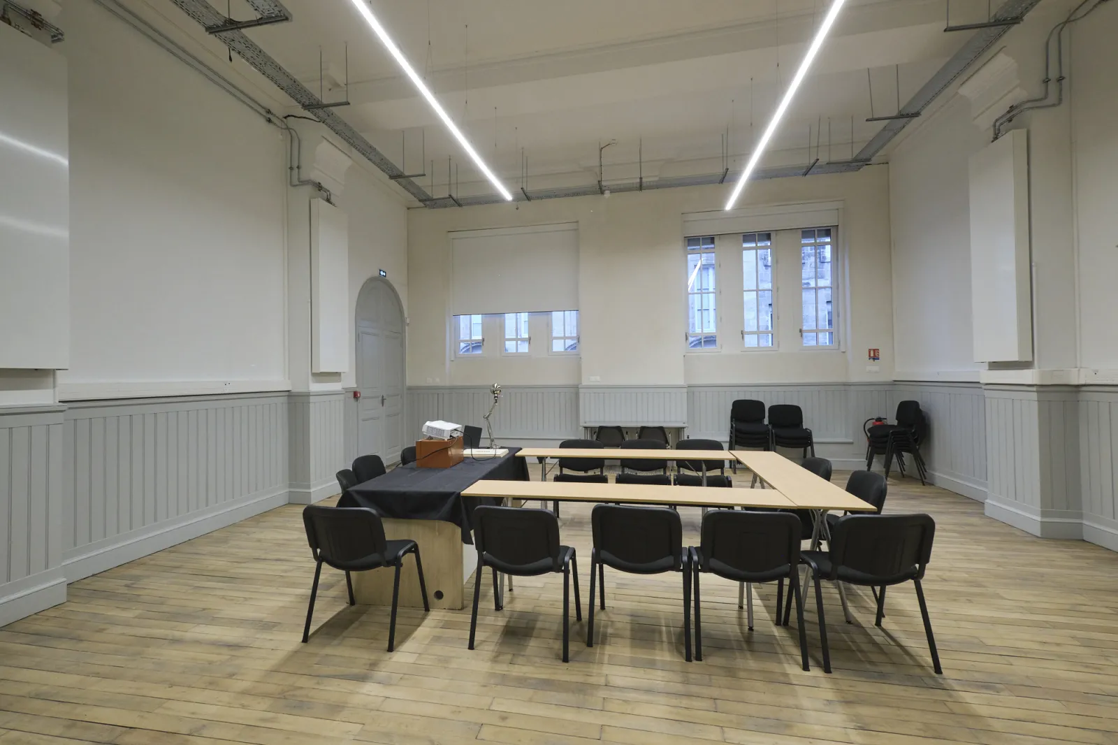 Modern conference room at ManuCo in Bordeaux with a large wooden table, black chairs arranged around it, white walls with wainscoting, bright LED ceiling lights, and tall windows providing natural light