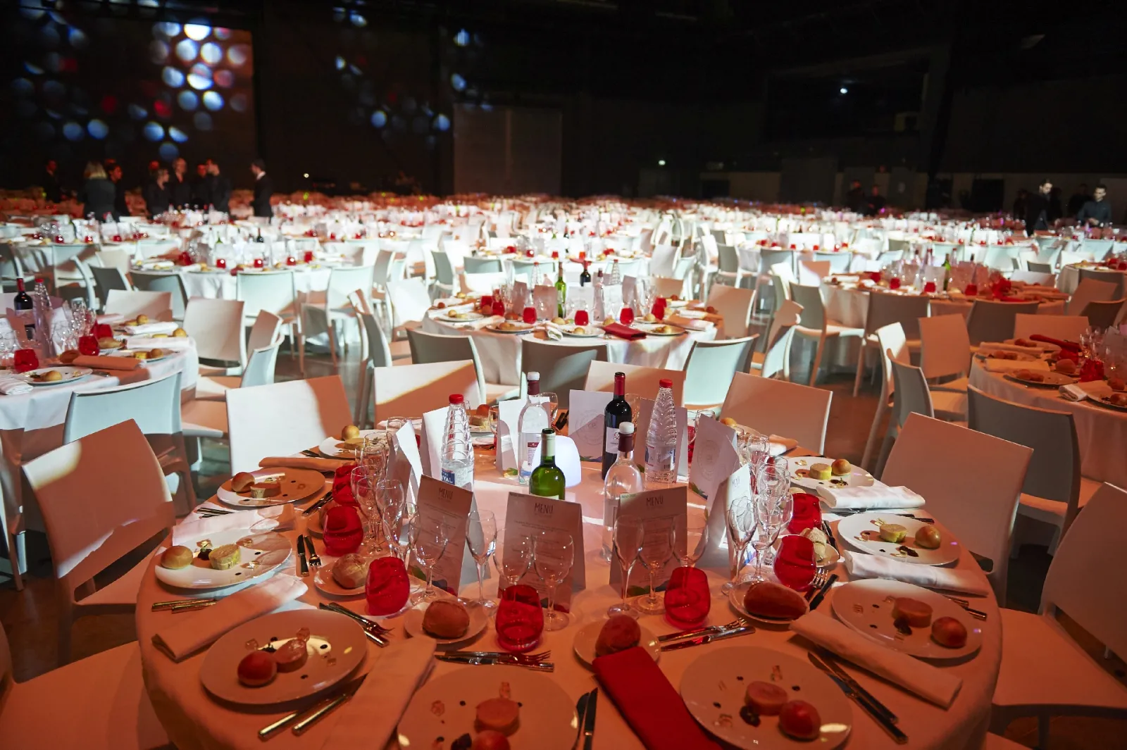 Large elegant banquet hall with hundreds of round tables set with orange tablecloths, white chairs, red candles, place settings, and wine glasses under dramatic blue lighting