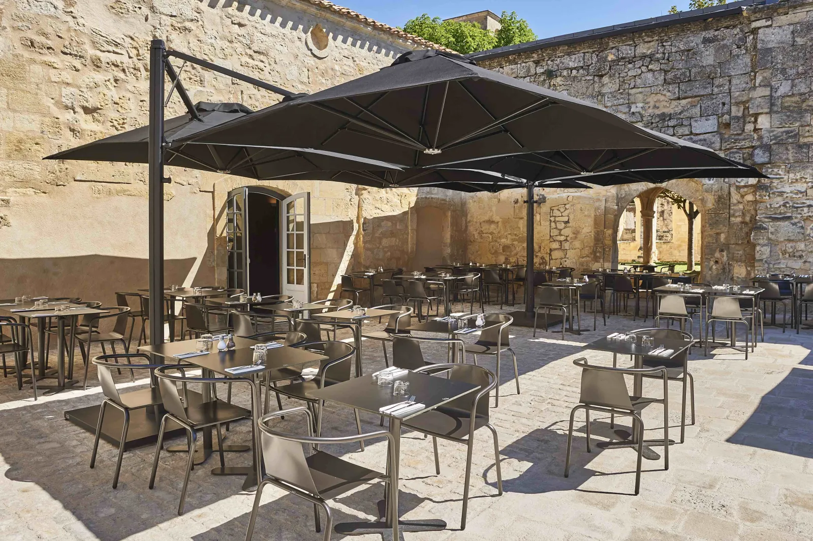 Outdoor terrace restaurant in a historic stone courtyard with black cantilever umbrellas, metal tables and chairs, and traditional architecture in the Cordeliers district of Bordeaux.