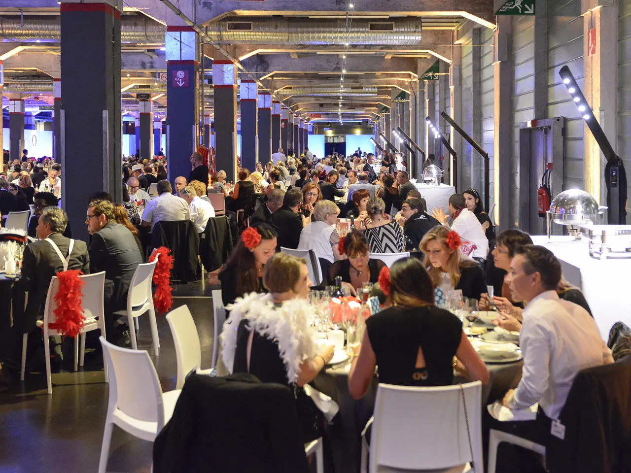 Large formal gala dinner event in a modern industrial venue with hundreds of guests seated at tables, decorated with red feather centerpieces, under ambient blue and gold lighting at Hangar 14 in Bordeaux.