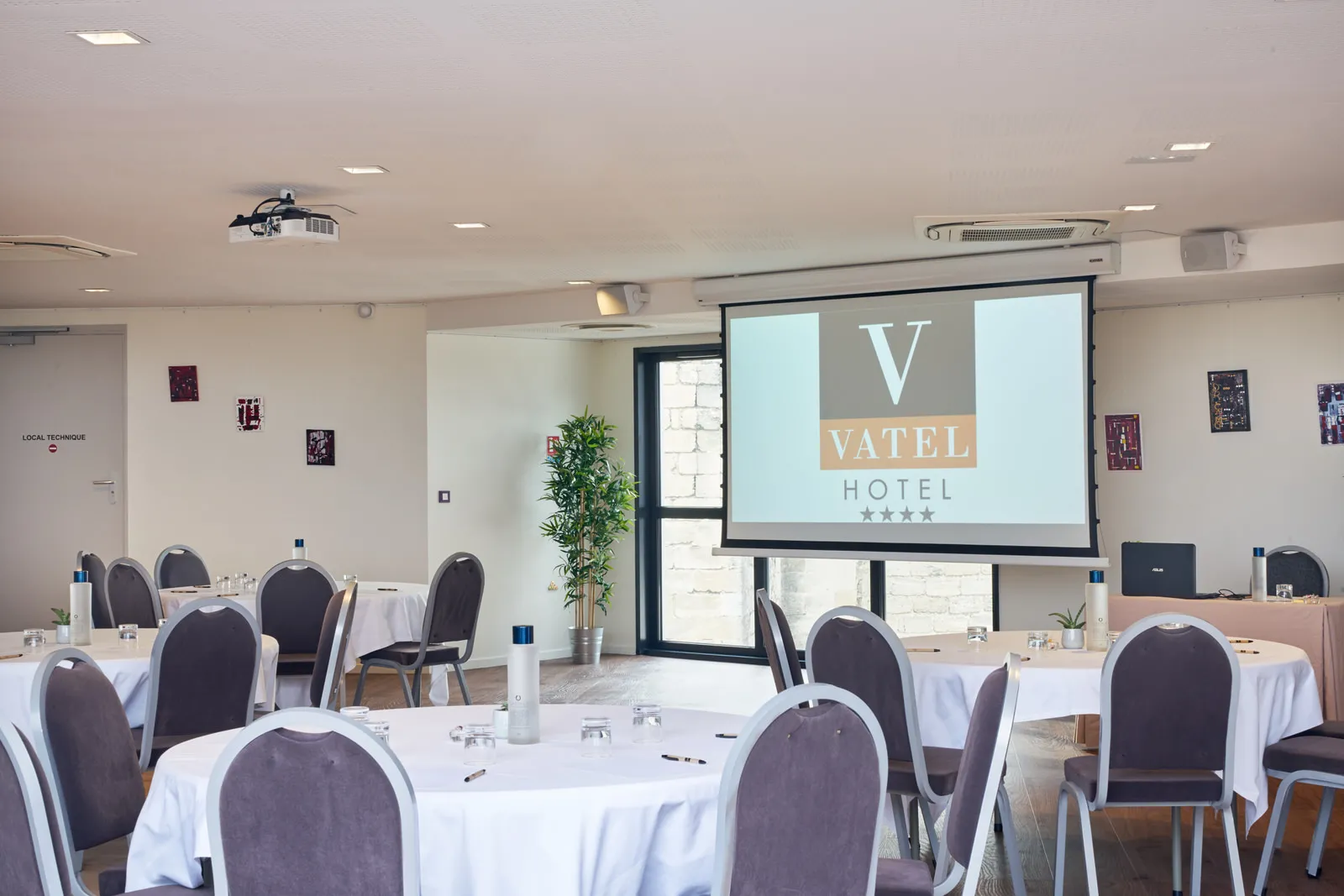 Conference room at Vatel Hotel in Bordeaux with round tables covered in white tablecloths, burgundy chairs, and a projection screen displaying the Vatel Hotel logo