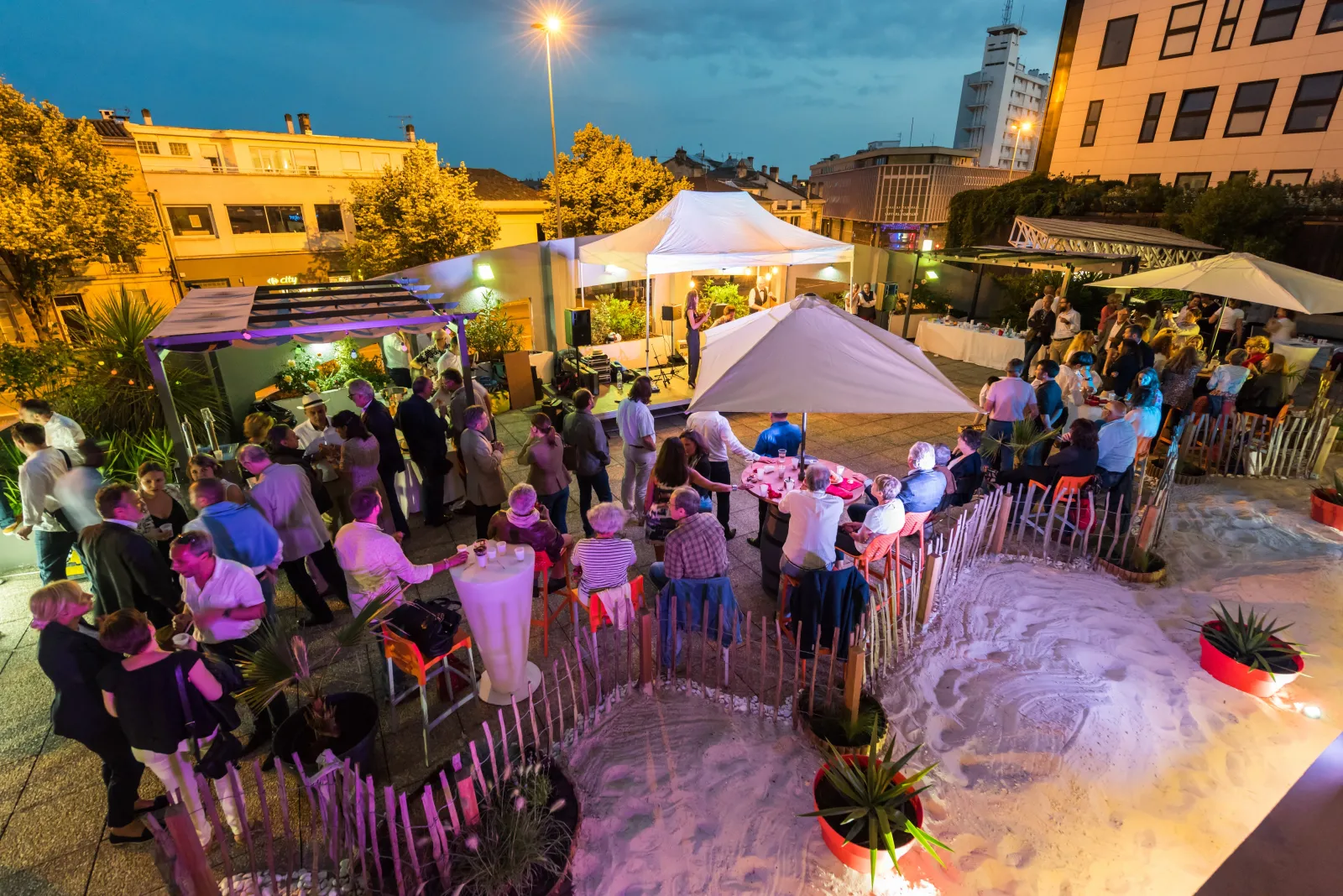 Evening rooftop event at Ibis Bordeaux Centre Mériadeck hotel with guests mingling around white marquees, tables, and lounge areas illuminated by purple ambient lighting, with city buildings visible in the background.