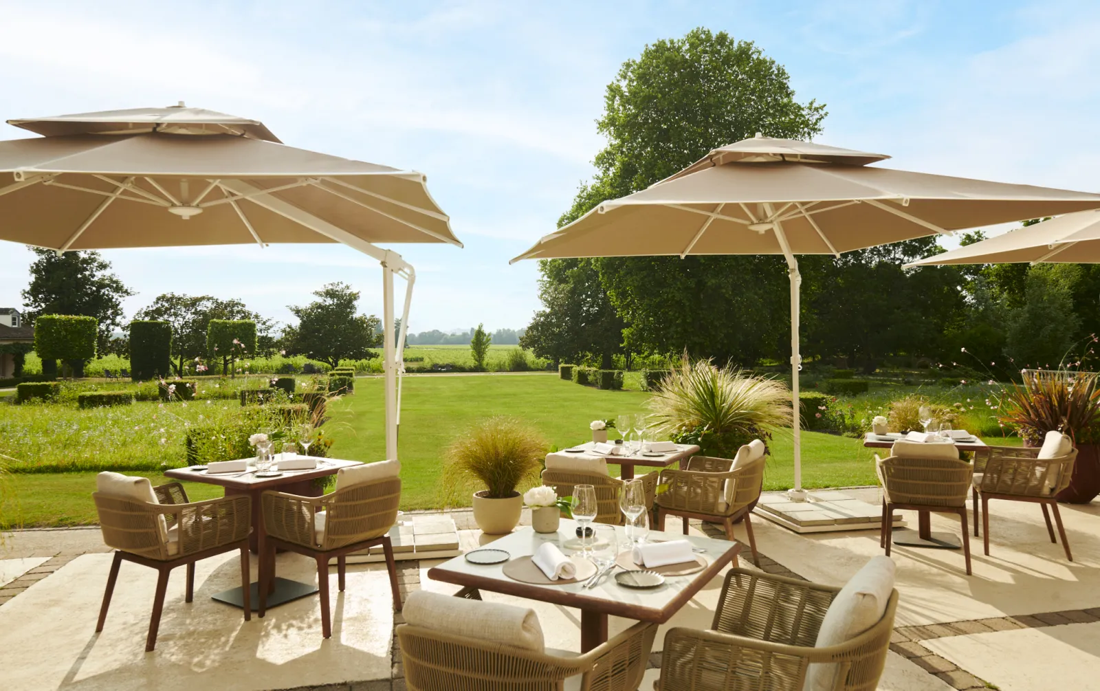 Elegant outdoor dining terrace at Château Hôtel Grand Barrail with beige cantilever umbrellas, woven chairs, and tables overlooking manicured gardens and green fields in Bordeaux