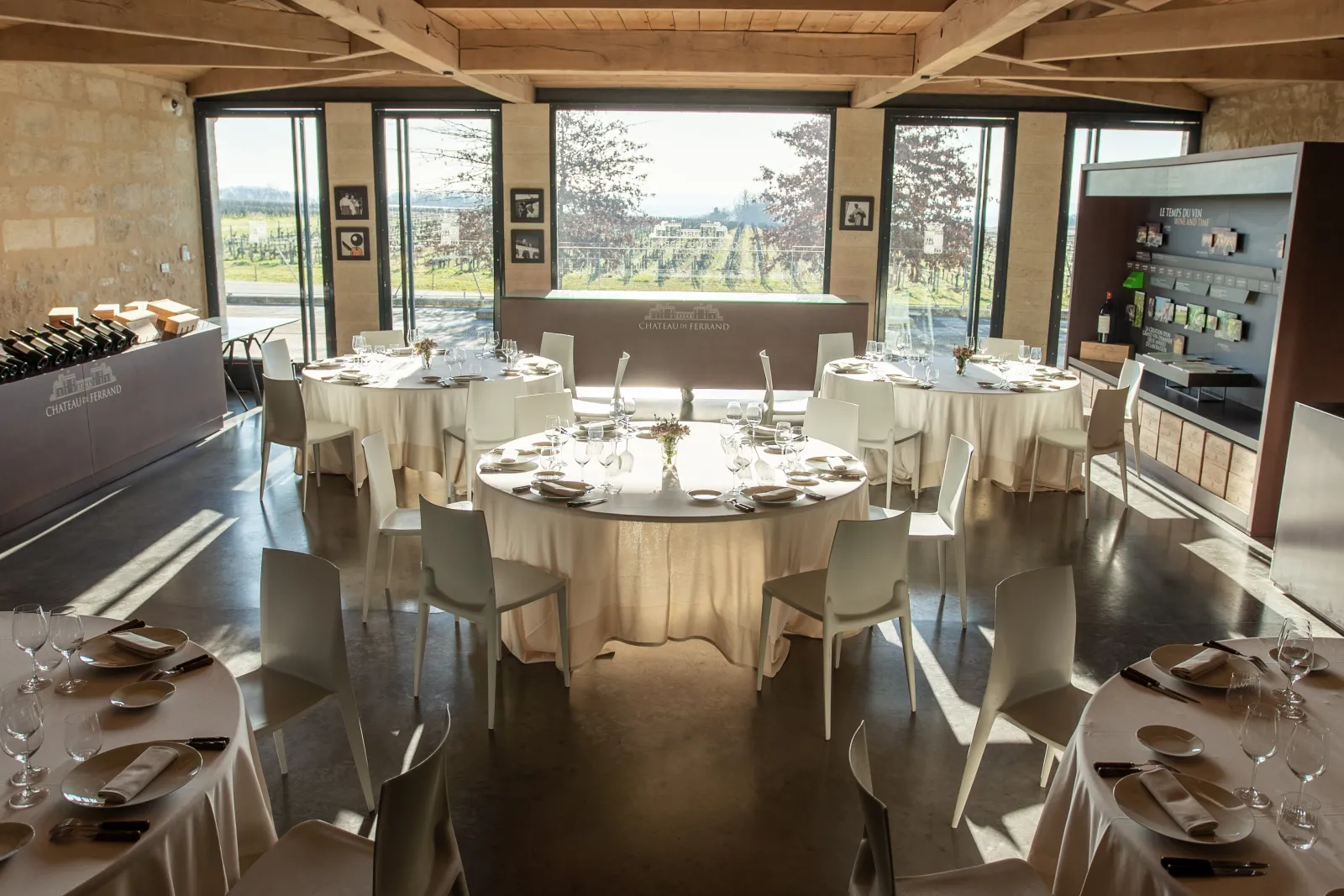 Elegant dining room at Château de Ferrand in Bordeaux with round tables set for fine dining, large windows overlooking vineyards, cream-colored tablecloths, and modern minimalist decor.