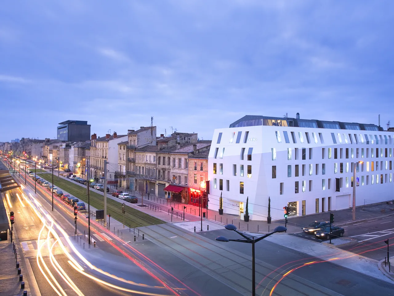 Modern white corner building of Seeko'o Hotel in Bordeaux at dusk, with historic architecture alongside, illuminated streets with light trails from traffic, and tram tracks visible