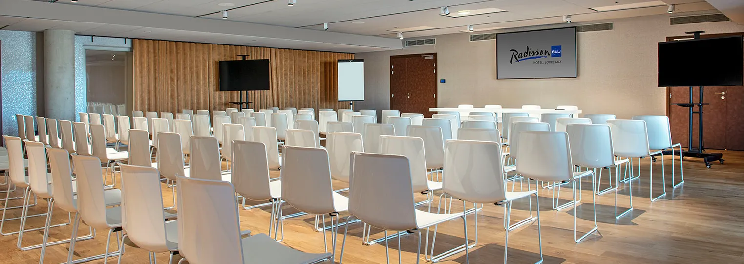 Modern conference room at Radisson Blu Hotel Bordeaux with white chairs arranged in rows, wooden wall paneling, projection screens, and professional audio-visual equipment.
