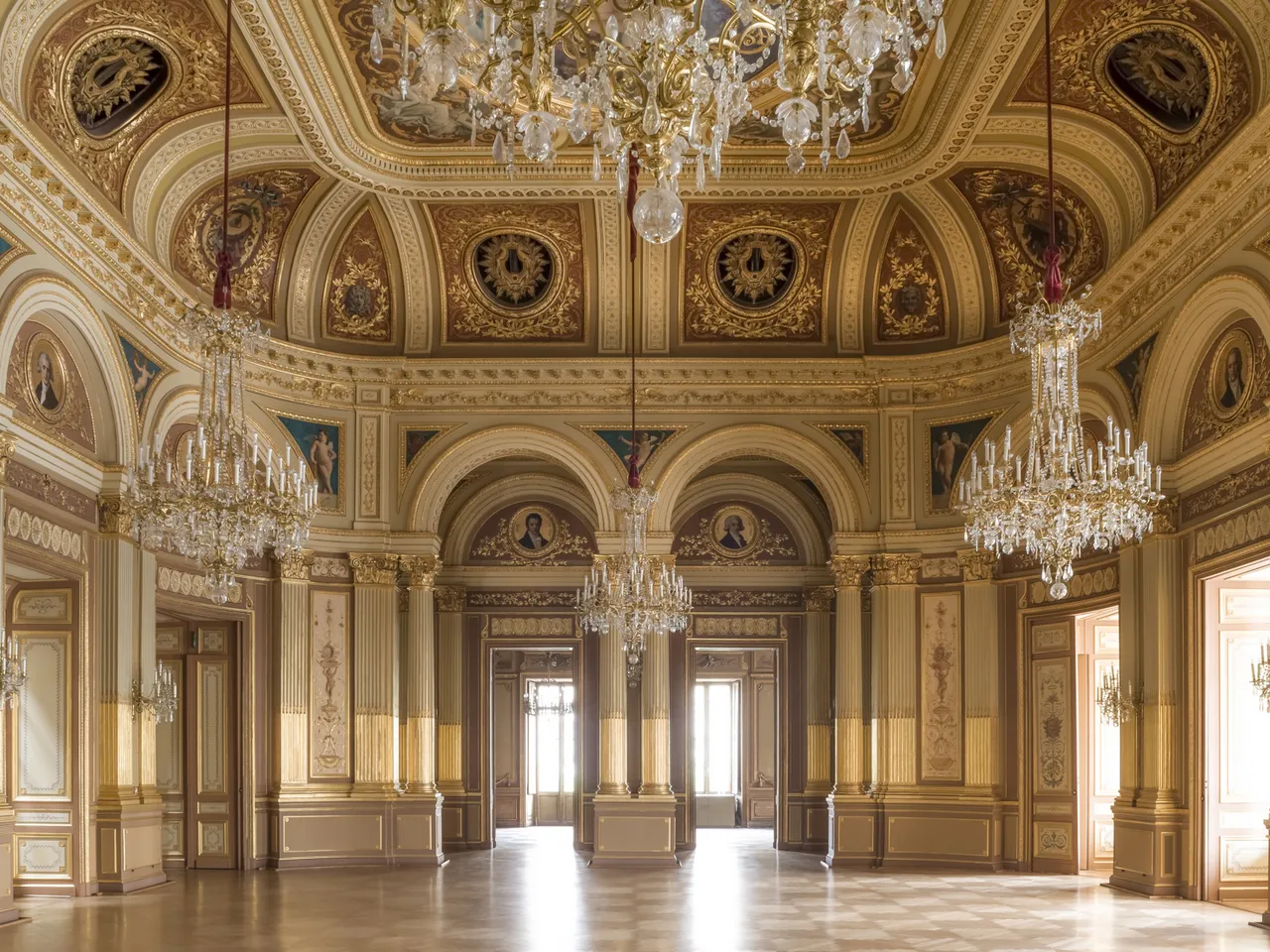 Grand ornate ballroom of the Bordeaux Grand Theatre featuring gilt decorative ceiling with frescoes, crystal chandeliers, fluted columns, and polished wooden floor