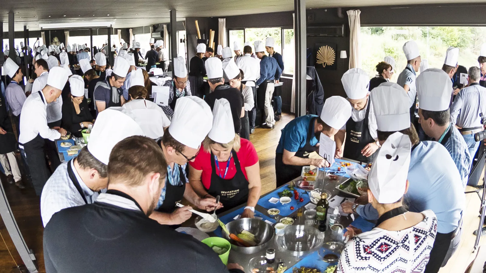 A large group of people wearing white chef's hats and aprons participate in a cooking class or culinary workshop in a modern venue with wooden flooring and panoramic windows overlooking greenery.