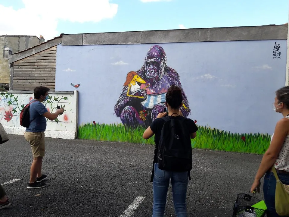 Street art mural in Bordeaux depicting a large purple gorilla holding a colorful bird, with green grass at the base, while visitors observe and photograph the artwork.
