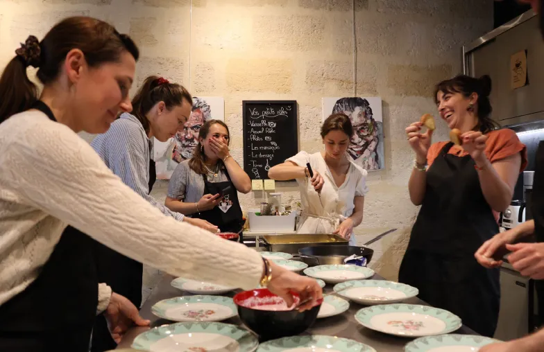 A group of people participating in a cooking class at La Cuisine de Camille in Bordeaux, plating dishes together at a counter with white plates and a black bowl.