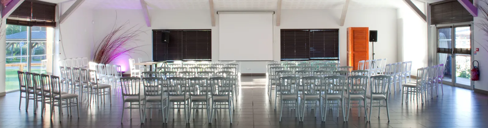 Modern event venue with gray chairs arranged in rows facing a central aisle, decorated with pink ambient lighting and decorative grasses, large windows overlooking green landscape, and minimalist interior design at Yelloh! Village Bordeaux Lac.