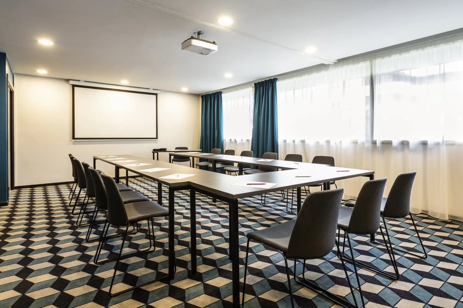 Modern conference room at Mercure Bordeaux Centre hotel featuring a large U-shaped table setup with black chairs, a projection screen on the wall, floor-to-ceiling windows with teal curtains, recessed ceiling lights, and a geometric patterned floor.