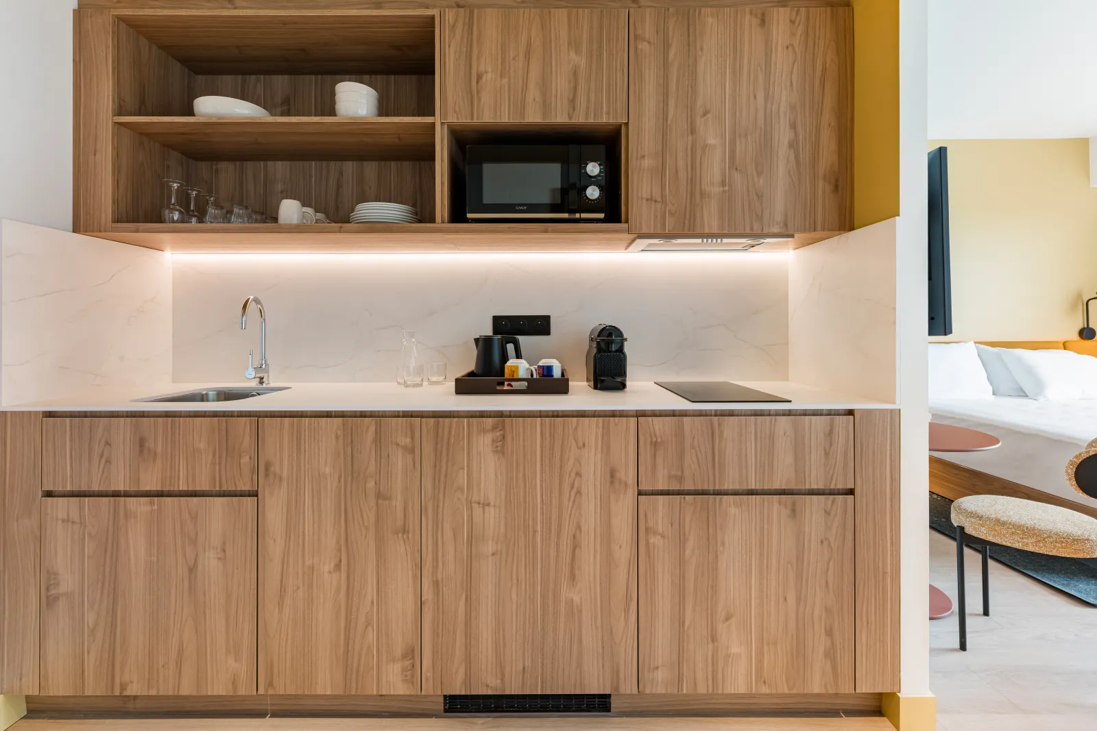 Modern kitchenette with wooden cabinetry, white countertop, integrated microwave, and ambient lighting in a hotel room at Hampton By Hilton Bordeaux Mérignac