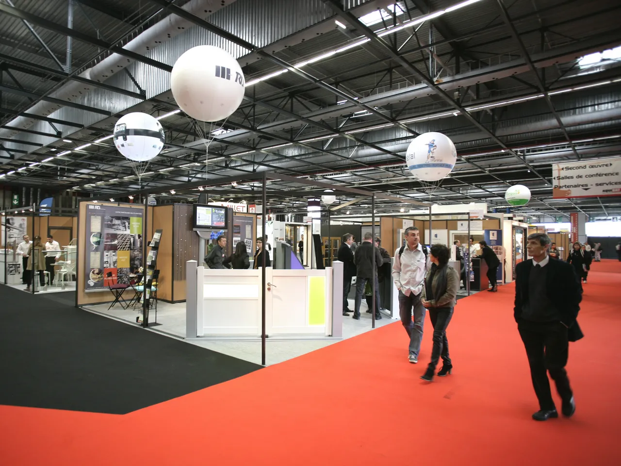 Interior view of the Bordeaux Exhibition Centre with visitors walking on a red carpet, modern booth displays with glass and wood elements, white spherical hanging lights, and industrial ceiling structure.