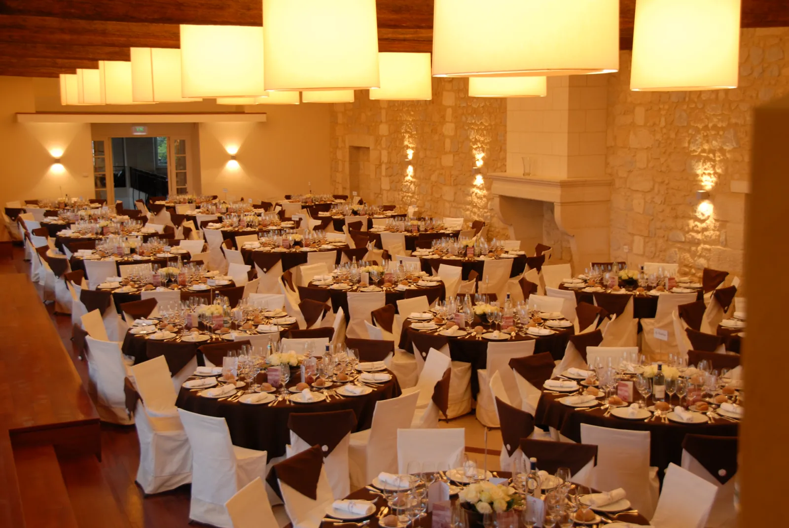 Elegant banquet hall at Château Léoville Poyferré in Bordeaux, featuring multiple round tables dressed with white linens, chocolate-colored chair sashes, place settings, and warm ambient lighting from ceiling fixtures.