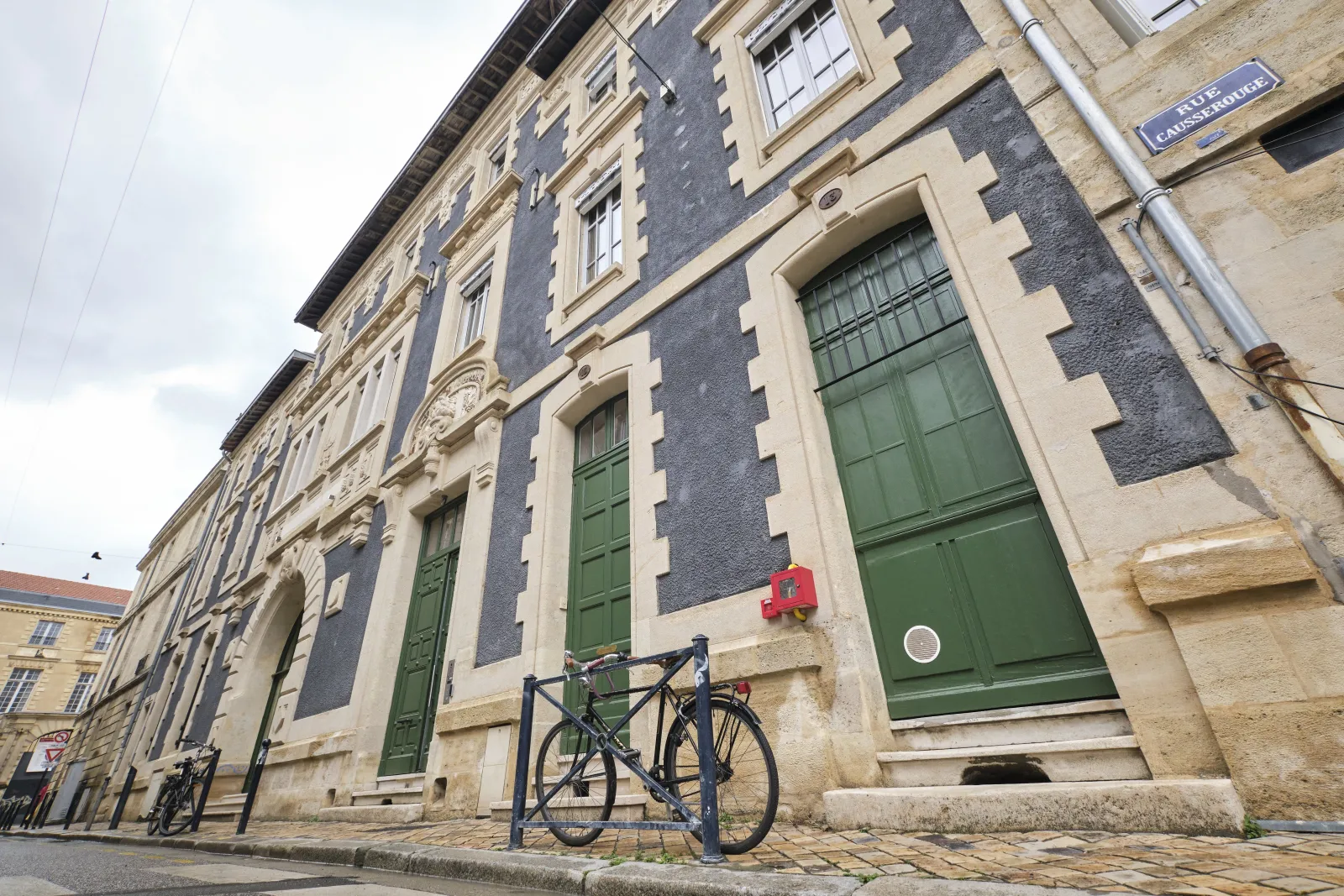Historic stone building with blue and cream striped facade, green doors, and a black road bike parked against a metal stand on the cobblestone street in Bordeaux