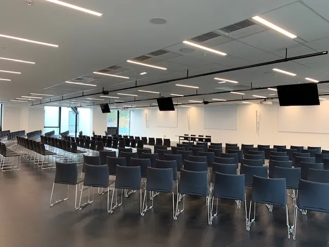 Large empty auditorium with rows of blue chairs, white walls, linear ceiling lights, projection screens, and windows at Palais 2 l'Atlantique in Bordeaux.