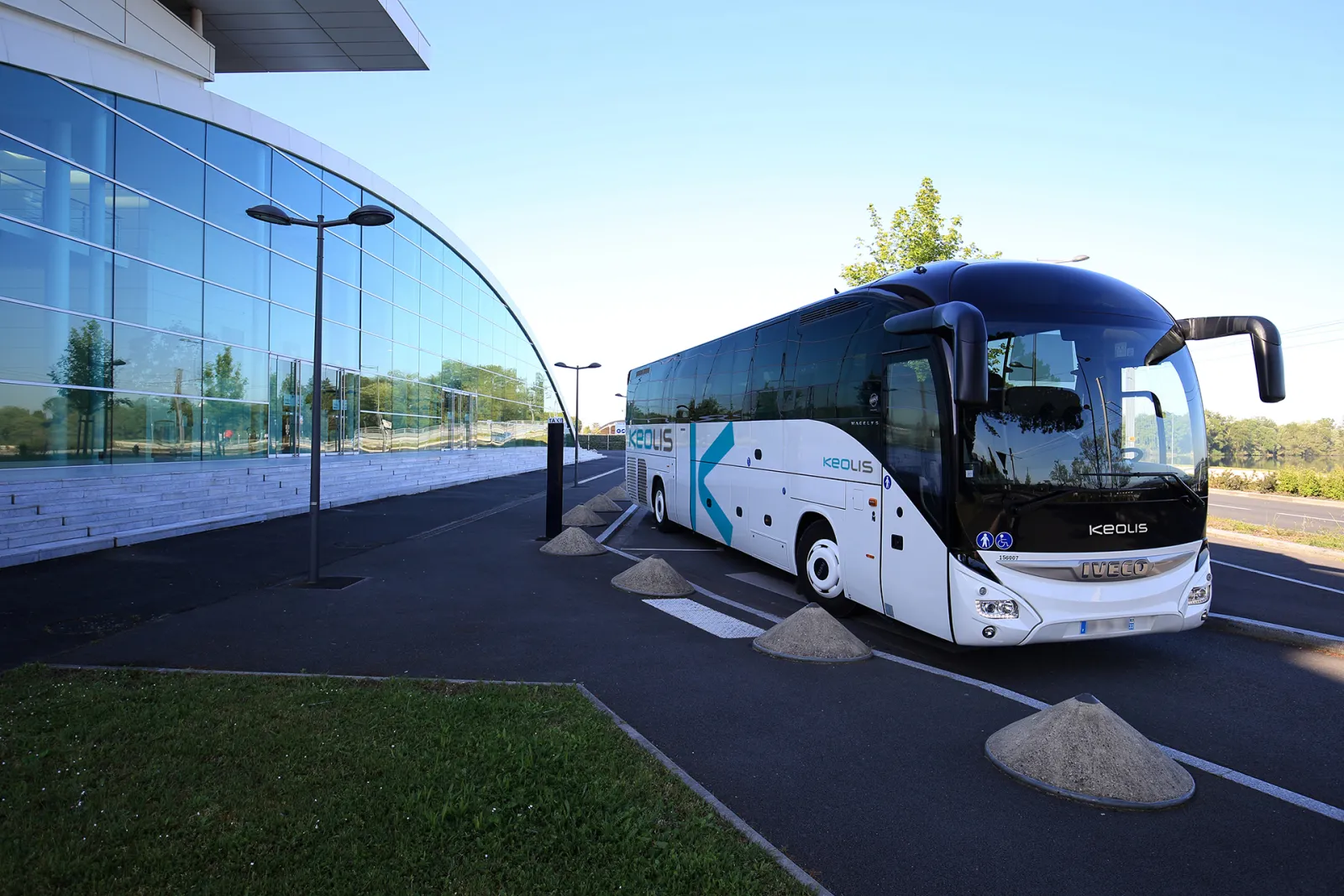 A white and blue Keolis coach bus parked in front of a modern glass building in Bordeaux, with street lamps and green spaces visible in the surroundings.