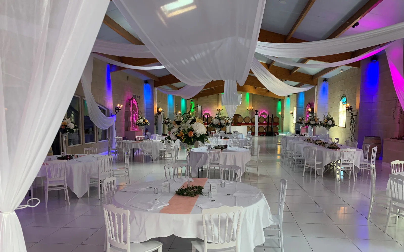 Elegant wedding reception hall at Château Lafitte in Bordeaux, featuring round tables with white tablecloths, white Chiavari chairs, draped white fabric ceiling decorations, colorful LED lighting in pink, blue, and green, and floral arrangements with whit