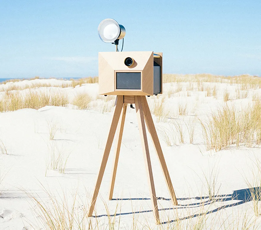 A wooden vintage-style camera on a tripod mounted on a sandy beach with dune grass and ocean in the background
