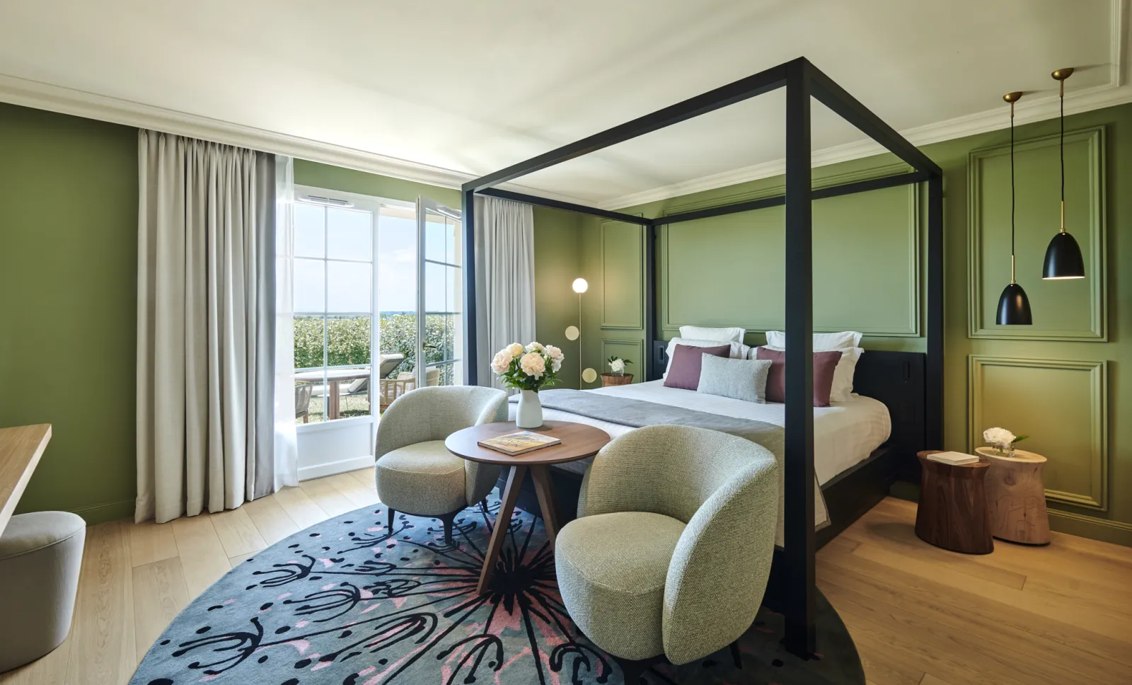 Luxurious hotel bedroom at Château Hôtel Grand Barrail in Bordeaux, featuring a black four-poster bed with white linens, sage green walls, contemporary seating area with cream armchairs, and panoramic windows overlooking the vineyard landscape.