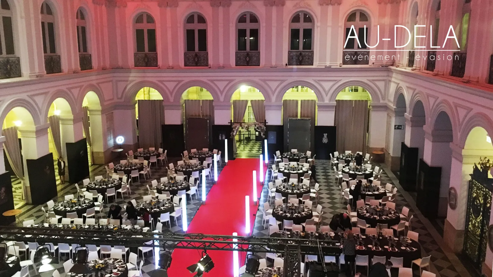 Aerial view of an elegant gala event in a pink-lit classical courtyard in Bordeaux, featuring multiple round dining tables, a red carpet with glowing edges, and arched galleries with ornate architectural details.