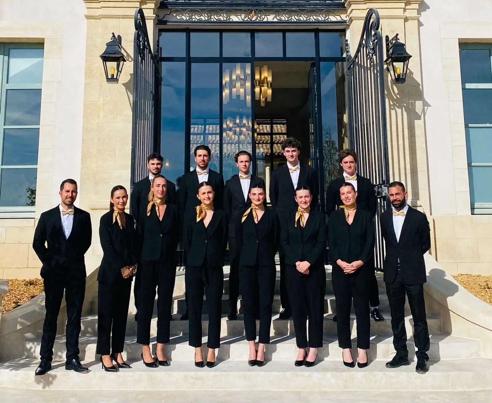 Professional team of 14 people in formal black suits and gold accessories posing in front of an elegant Bordeaux mansion entrance with black iron doors and classical architecture.