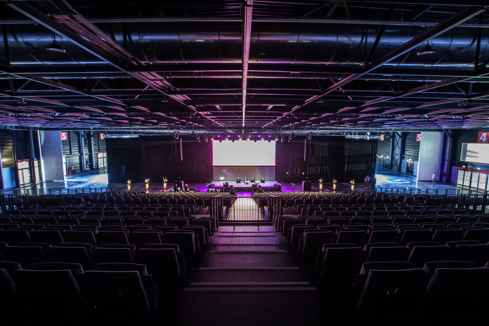 Large empty auditorium with black seats facing a stage illuminated by purple and blue neon lights, industrial ceiling with exposed ductwork, and a projection screen at Palais 2 l'Atlantique in Bordeaux.