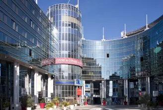 Modern architectural complex featuring a cylindrical glass tower with white framework, surrounded by curved blue-tinted buildings. The central plaza displays signage for 'Centre de Congrès' with decorative red planters and landscaping under clear blue sky