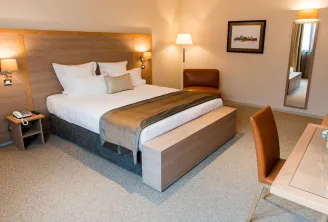 Modern hotel bedroom at Vatel Bordeaux featuring a king-size bed with white linens, wooden headboard with integrated lighting, bench at the foot of the bed, leather armchair, desk area, and ensuite bathroom visible through an open door.