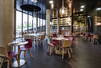 Modern restaurant interior with wooden chairs and burgundy seats arranged around small tables, featuring a sophisticated bar with extensive bottle selection, pendant lighting, and floor-to-ceiling windows overlooking greenery.